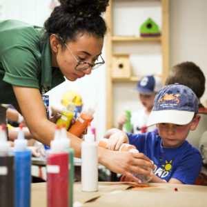 Reina Argüello, PTC Craft Center Staff, helps Gus Bower with making a sand sculpture using multiple different colors off sand, and a glass container to hold the sand.